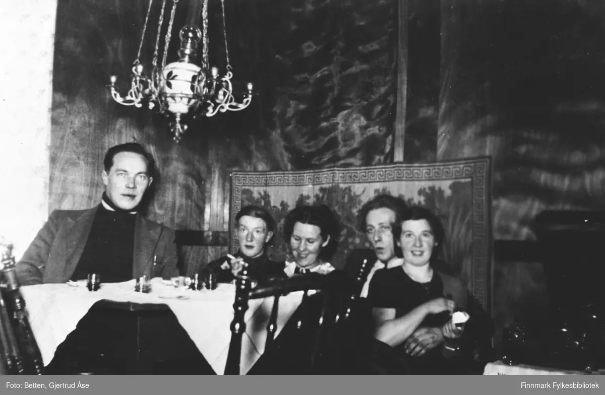 To kvinner og tre menn sitter i stua på hytta hos Ingeborg. Fra venstre: Abraham Mikkelsen, Nils Betten, Ingeborg Moan, Otto Pleym og Gjerdrud Betten. Det henger en oljelampe over bordet, bordet er dekt med en hvit duk. På bordet står det små likørglasser men noe oppi.  Under bildet på albumet står det ' I kosekroken hos Ingeborg'.