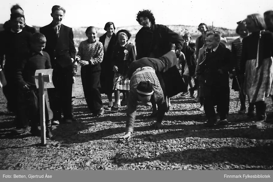 17.mai feiring ved Seida skole i 1956.   Barna og voksne leker ved skolen. En kvinne kjører trillebår med en gutt. Alle ser til å ha det artig. Dagen er solig og fin.