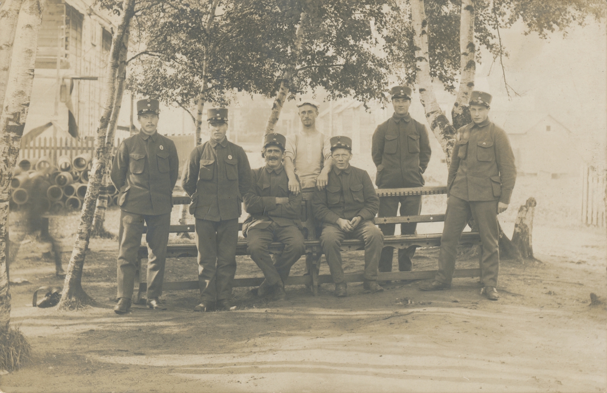 Gruppefotografi av menn i militæruniform og en i sivil ved en benk. Flere hus i bakgrunnen.