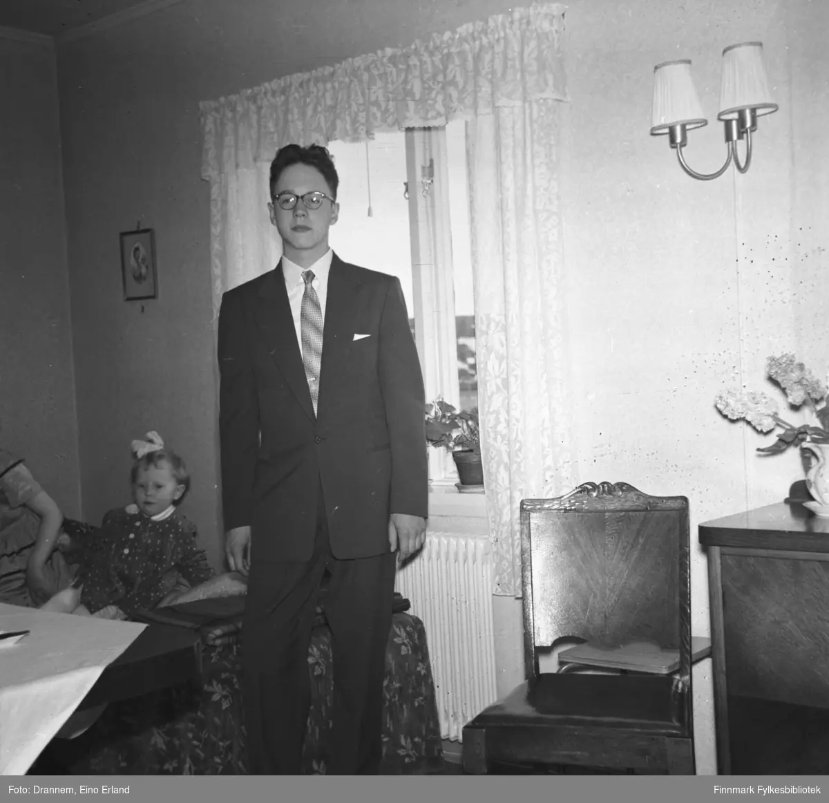 Arvid Sørensen, konfirmant ca. 1950, i stua til familien Drannem i Hammerfest på 17. mai. Grete Sørensen sitter på sofaen i bakgrunn.