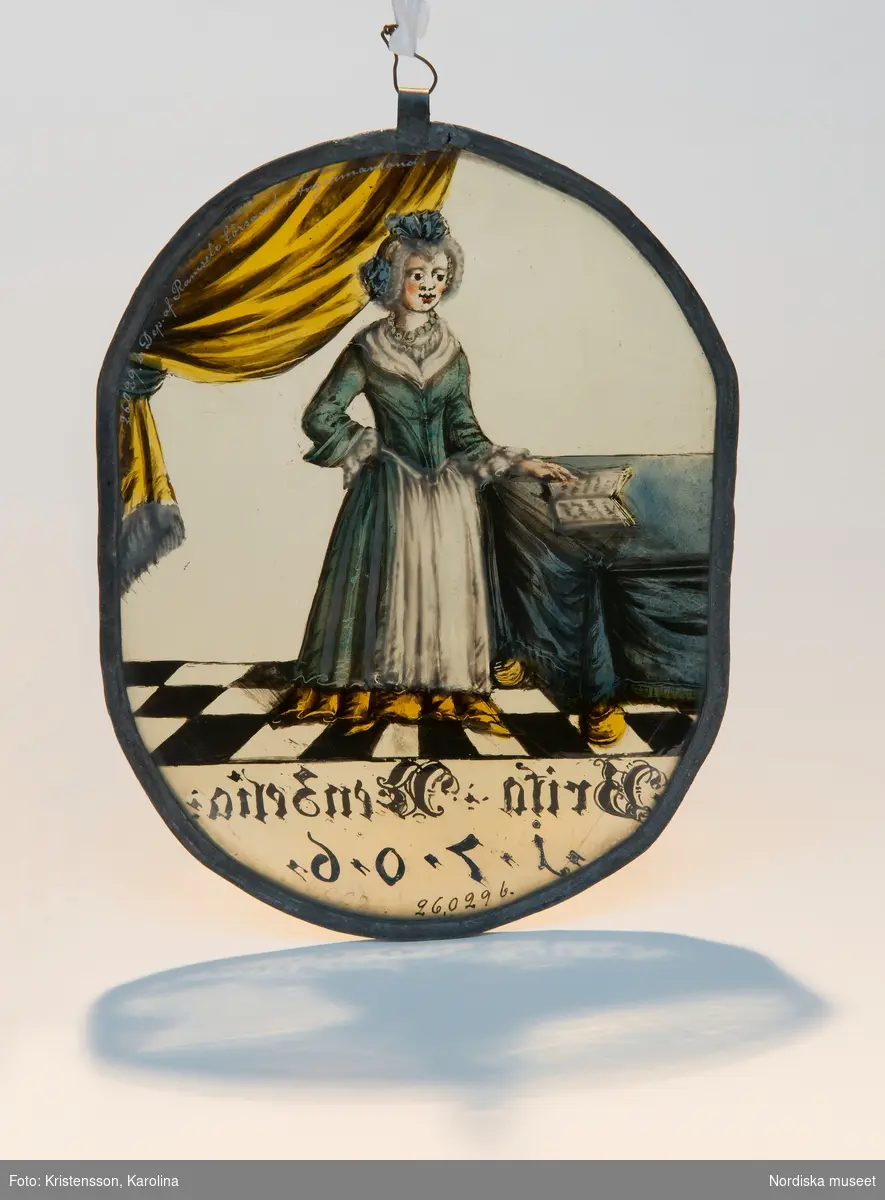 Fru Brita Nenzelius stående på ett rutat golv. Till vänster ett täckt bord med en bok, till höger ett draperi. Hon bär en blå klänning med vita isättningar, runt halsen ett halsband. Text: "Brita Nenzelia 1706".