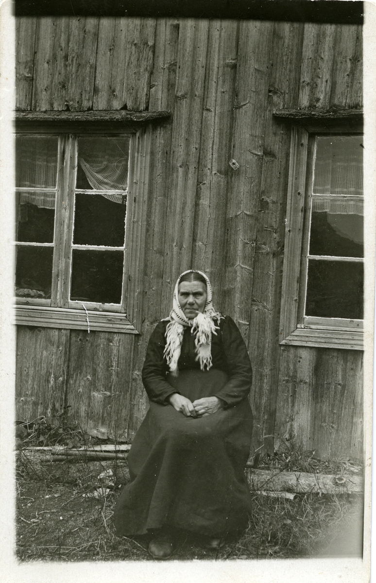 Portrett av en kvinne sittende foran en husvegg. Kvinnen er iført kjole og hun har et skaut på hodet.