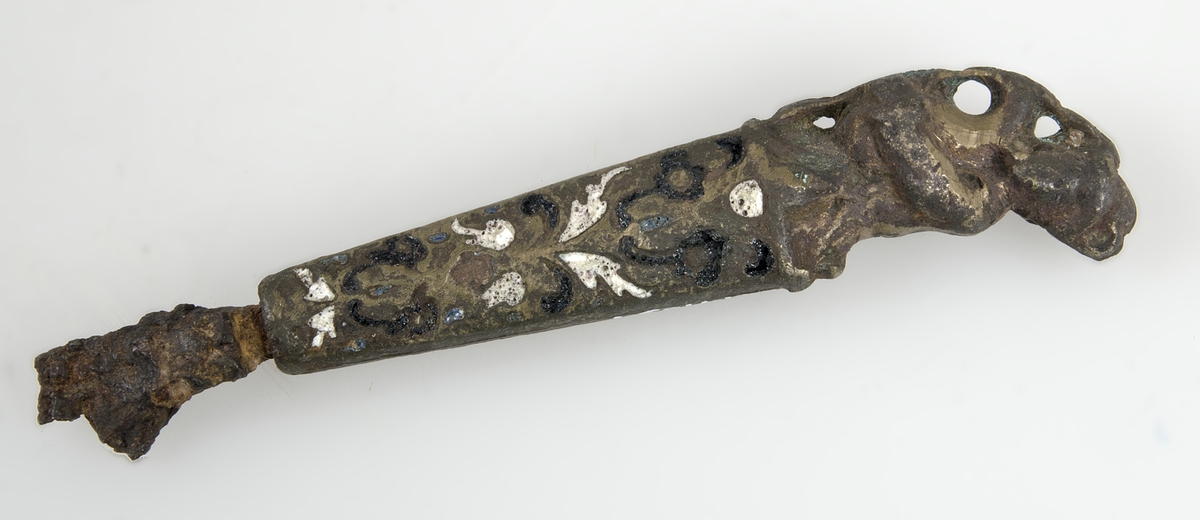 Knivskaft av brons med emaljinläggningar i vitt och mörkblått. Skaftet är längst upp format som en man som spelar säckpipa. Av knivbladet finns endast rester kvar.
Märkt "EC.603."