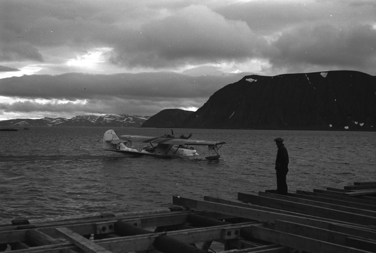 En flybåt, muligens en Catalina, takser ute på fjorden ved Honningsvåg. Foran på bildet er en kai under byggning. En person står på land og betrakter det hele. 

Flybåt er et fly som starter og lander på sjøen. En flybåt skiller seg fra et sjøfly ved at selve skroget flyter på vannet, skroget er dermed formet som på en båt. Eksempler på flybåter er for eksempel Short Sandringham og Dornier Do X. Flybåtene var svært populære før nettet av flyplasser var skikkelig utbygd, og egnet seg bedre for langdistanseflygninger enn samtidige landbaserte passasjerfly.
Arkitekt Ola Hanche-Olsen arbeidet ved Brente Steders Reguleringskontor i 1946. Hovedadministrasjon for gjenreisning av Nord-Troms og Finnmark ble lagt til Harstad og fikk navnet Finnmark kontoret. Landsdelen Nord-Troms og Finnmark blev oppdelt i syv distrikt med hver sin administrasjon. Honningsvåg, distrikt IV, skulle betjene Nordkapp, Lebesby, Porsanger og Karasjok kommune.

Ola Hanche-Olsen har tatt bildene. Han var født 13. mars 1920 i Borre, død 11. februar 1998 i Gjettum. Han var både arkitekt og barnebokforfatter. Han hadde artium fra 1939, arkitekteksamen fra NTH 1946 og arbeidet deretter ved Finnmarkskontoret 1946–48 før han etablerte egen arkitektpraksis. Han debuterte som barnebokforfatter i 1974 med lettlest-boka "Knut og sjørøverne", og skrev i alt 12 bøker. Han var XU-agent 1944-45, og var også en aktiv fjellklatrer og friluftsmann. Ola var gift med Solveig Hanche-Olsen (f. Falkenberg); de fikk 3 barn, blant dem matematikeren Harald Hanche-Olsen.


