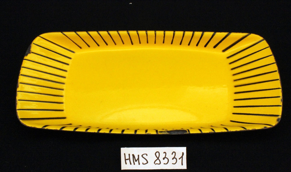 Rektangulært brett i emaljert emalje med avrundete hjørner, gul bunnemalje med sorte striper. 
Designet av Grete Prytz Kittelsen for Cathrineholm fabrikk i Halden. 