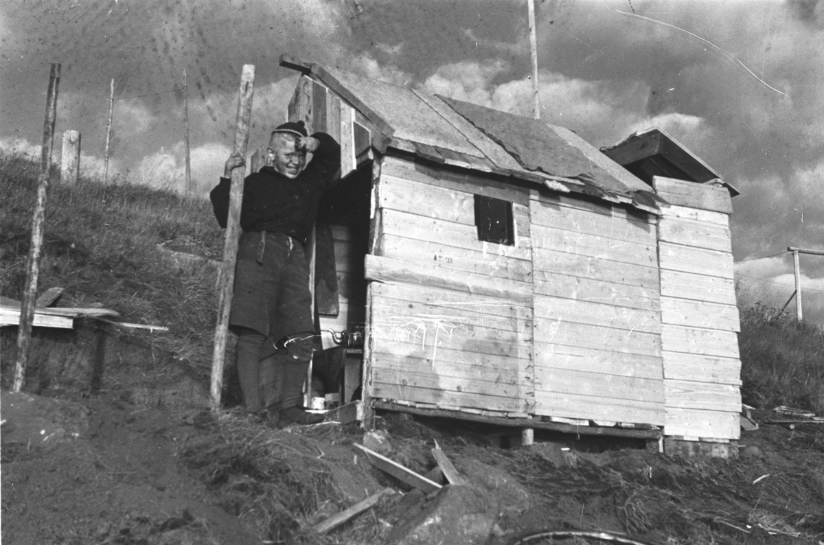 En gutt står ved siden av en lekehytte som sannsynligvis er bygget av materialrester fra gjenreisningsbygg i Honningsvåg etter andre verdenskrig.

Arkitekt Ola Hanche-Olsen arbeidet ved Brente Steders Reguleringskontor i 1946. Hovedadministrasjon for gjenreisning av Nord-Troms og Finnmark ble lagt til Harstad og fikk navnet Finnmark kontoret. Landsdelen Nord-Troms og Finnmark blev oppdelt i syv distrikt med hver sin administrasjon. Honningsvåg, distrikt IV, skulle betjene Nordkapp, Lebesby, Porsanger og Karasjok kommune.

Ola Hanche-Olsen har tatt bildene. Han var født 13. mars 1920 i Borre, død 11. februar 1998 i Gjettum. Han var både arkitekt og barnebokforfatter. Han hadde artium fra 1939, arkitekteksamen fra NTH 1946 og arbeidet deretter ved Finnmarkskontoret 1946–48 før han etablerte egen arkitektpraksis. Han debuterte som barnebokforfatter i 1974 med lettlest-boka "Knut og sjørøverne", og skrev i alt 12 bøker. Han var XU-agent 1944-45, og var også en aktiv fjellklatrer og friluftsmann. Ola var gift med Solveig Hanche-Olsen (f. Falkenberg); de fikk 3 barn, blant dem matematikeren Harald Hanche-Olsen.

