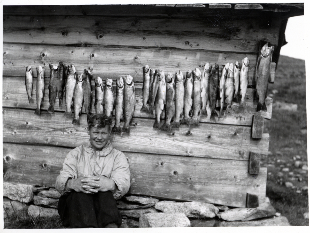 Portrett av en ung mann sittende foran en husvegg. Bak seg henger det en lang rekke med fisk.