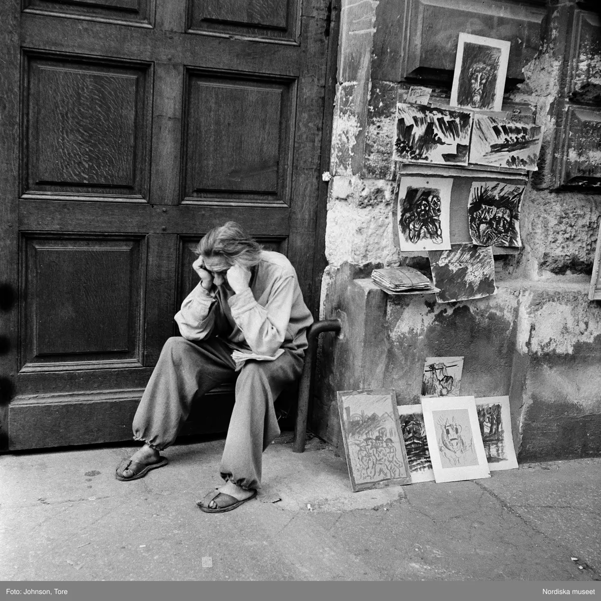 Paris. En man sitter med ansiktet i händerna. Målningar/skisser till salu uppsatta på husfasaden bredvid.