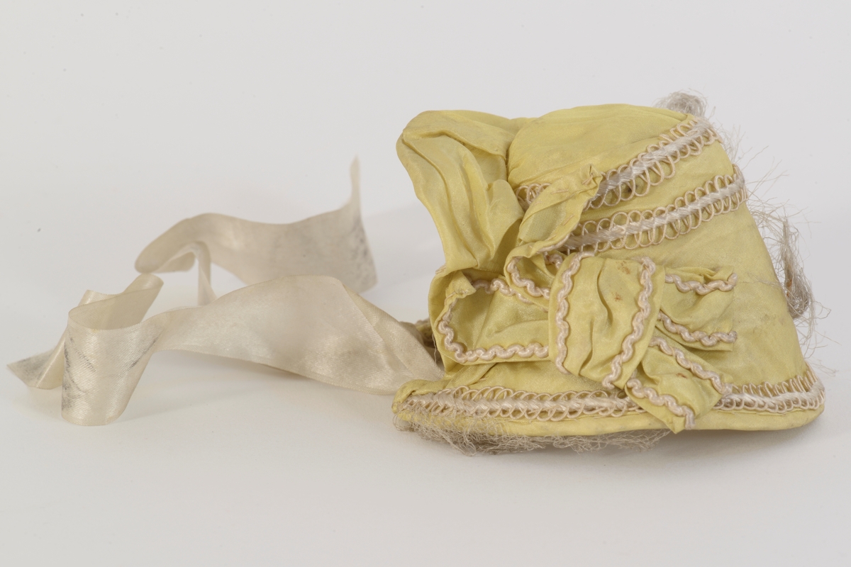 Dukkekyse i (oppr) gul silke med pyntebånd. Avstivet og pyntet med tyllkant og bånd. Foret med silke. Knytebånd i silke.