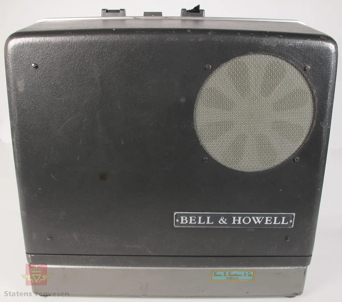 BELL & HOWELL MODEL 642. 16 mm filmfremviser i mørk grå koffert av glassfiber, med defekt bærehåndtak. Den ene delen av kofferten har høyttaler av merket Goodmans, ledning for tilkobling av høyttaler og to klistremerker med påskrifter. Filmfreviseren har eget panel for justering av lyden. Bak på kofferten er det to skiltplater med innskrifter, MODEL 642 SK SERIAL No. K418 220V-1100 W - 50 ~ MADE IN ENGLAND og HANS H. KNUTSEN & CO. OSLO TYPE 642 No 418 P.VOLT 200/250 50 ~ S.VOLT AMP. 1200W. Veske, strømledning, ekstra lyspære og to tomme filmruller følger med filmfremviseren.