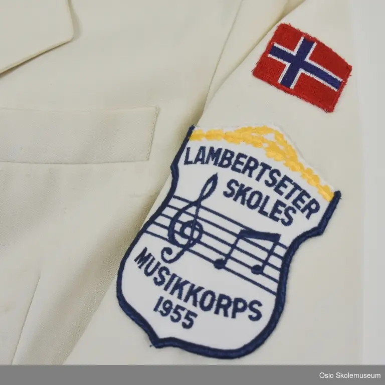 Beige jakke fra Lambertseter skoles musikkorps til barn. Jakken har korpsets emblem og Norges flagg på ermet. Gule tråddekorasjon på skuldrene og knapper i plast.