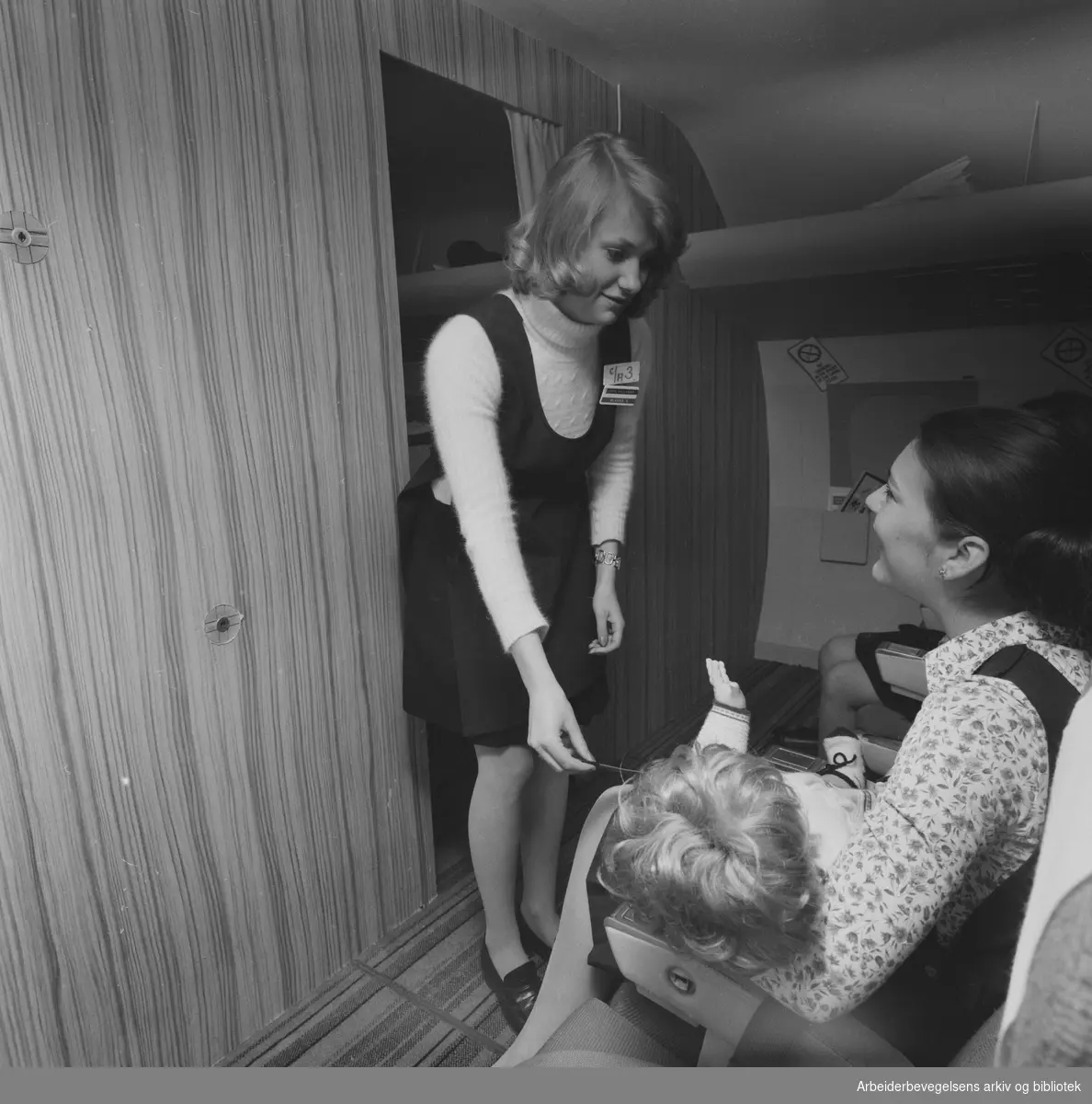 Kurs for flyvertinner i SAS på Park Hotel i Sandefjord. Undervisningen finner sted i en tro kopi av kabinen på en DC-9 i kjelleren på hotellet. "Mamma" Christina Stineberg får hjelp av vertinneelev Lotta Hillander. April 1973.