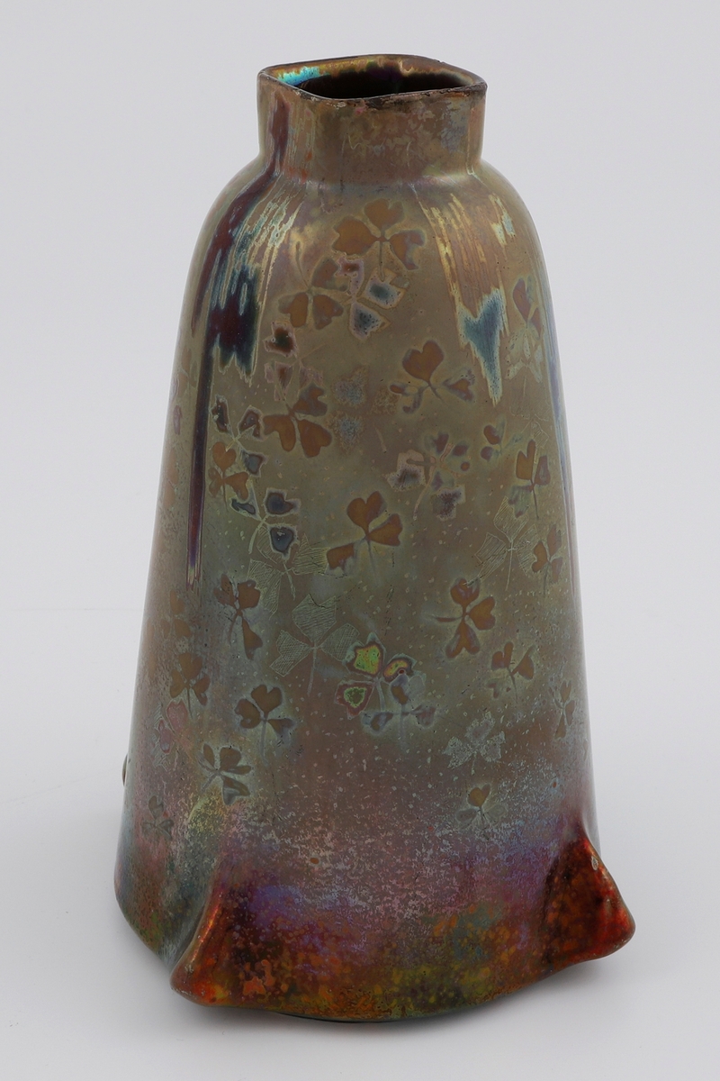 Konisk vase i glasert steingods med lusterglasur. Nedre del av vasen er markert med fire utstående "ører". Formen smalner gradvis oppover, og avsluttes med en firkantet munningsrand med avrundede hjørner. Lusterglasuren har en metallisk, regnbueskimrende virkning, som veklser mellom røde, grønne, gule, brune og rød-lilla fargenyanser. På deler av vasen er det fremstilt silhuetter av trekløver.