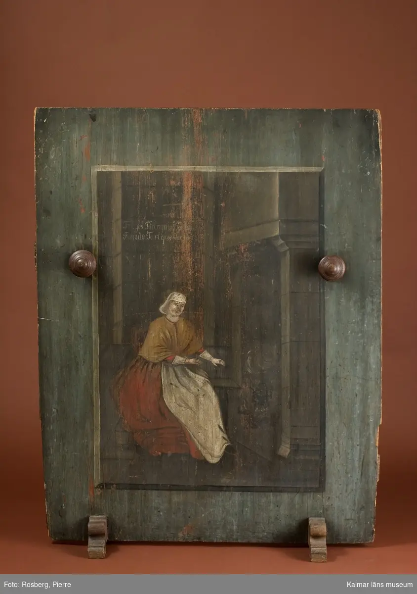 Porträtt av kvinna, helfigur, interiörmotiv, kvinnan sitter framför spisen med eld och kokkärl.