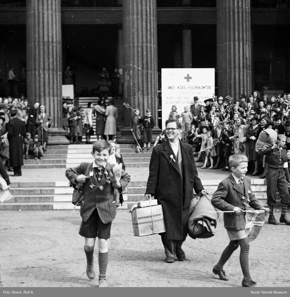 Far med to sønner med oppakning går over Universitetsplassen, ved Røde Kors hjelpekontor, folkemengde i bakgrunnen, Oslo, mai 1945,