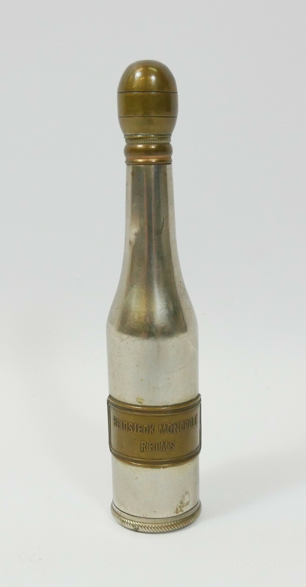 Liten flaske i blank metall med kobberfarget skrukork og plankett. 

Påskrift: 
HEIDSIECK MONOPOLE // REIMS 