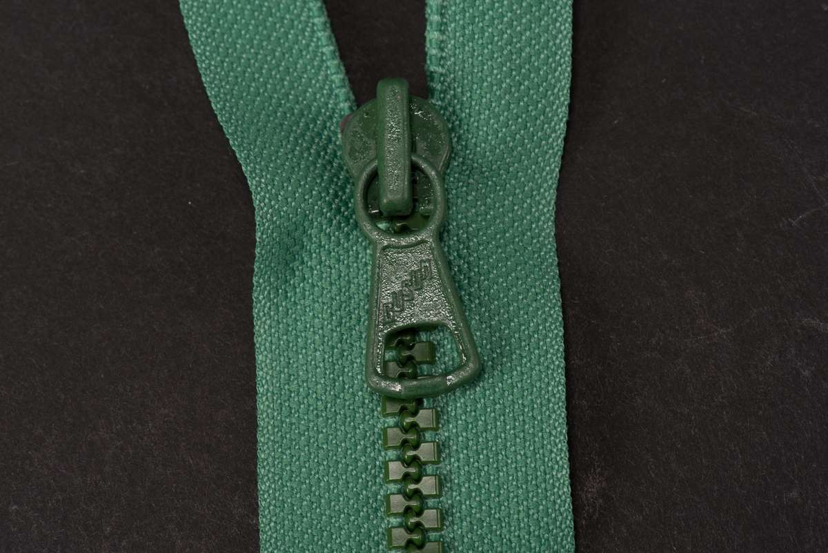 Grön dragkedja med textilband, häktor av plast och löpare och dragtapp av metall. På dragtappen inpräglat GUSUM.