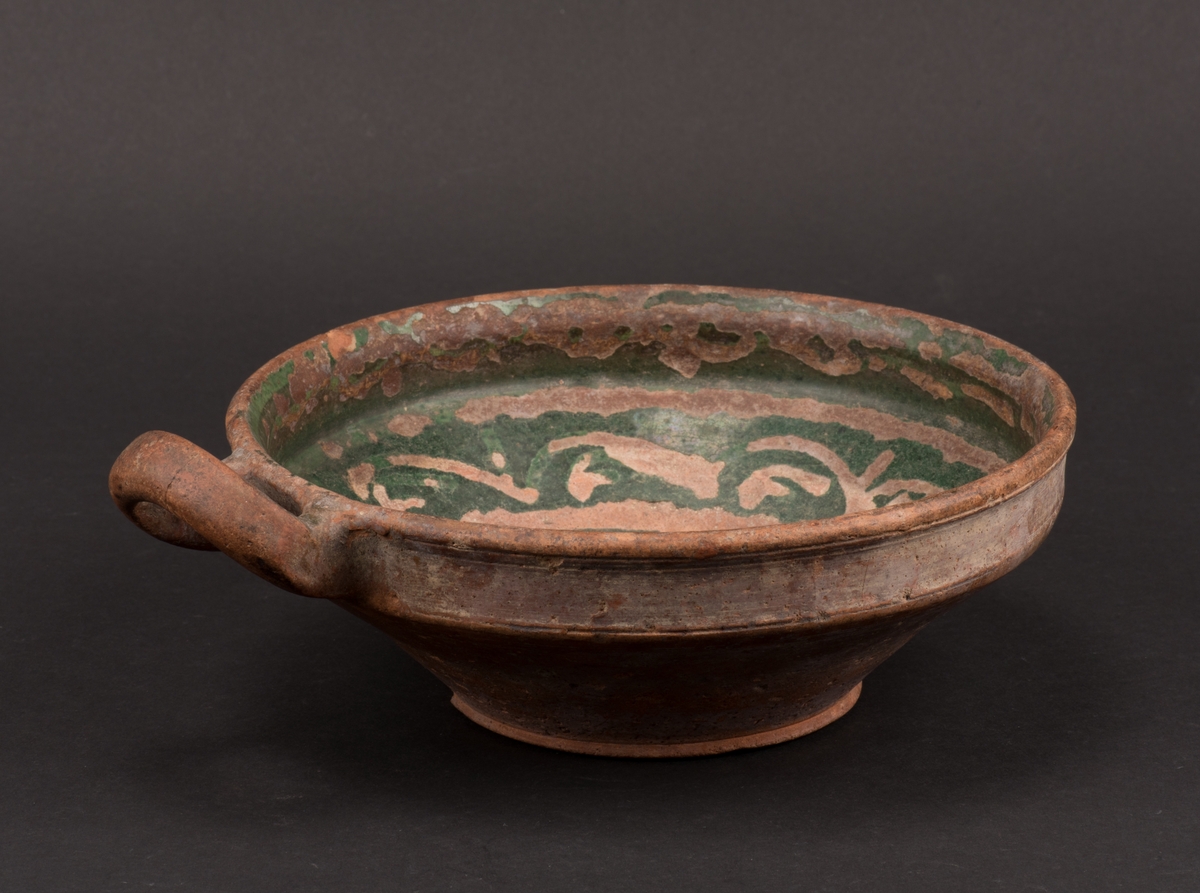 Rund, drejad skål av bränd lera. Konisk form med rak övre kant där skålen är försedd med ett öra. Grön glasering inuti skålen, med ränder och vågformat mönster samt årtalet 1830.