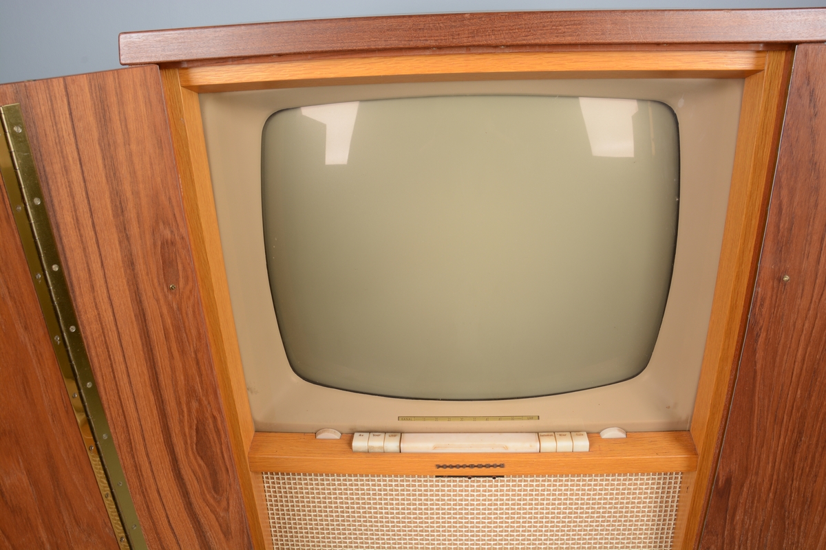 TV/fjernsyn montert i kabinett med dører for skjuling av fjernsynet når det ikkje var i bruk. i skåpet er og montert to høgtalarar. Beteningsknappar, antenne og elektrisk kabel. Kabinettet/skåpet har rektangulær form.