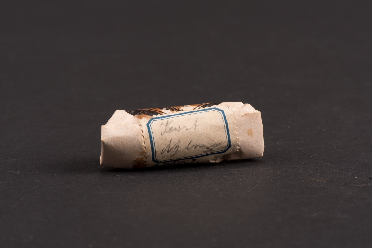 Rund begravningskaramell invikt i vitt papper. Dekorerad med en tryckt etikett som visar en gravsten med urna flankerad av fyra granar. 
På baksidan en etikett med ett namn "Kurt Nyborg".