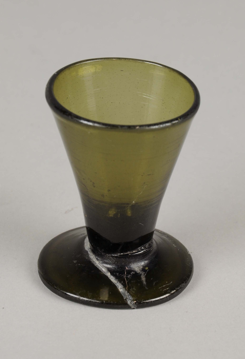 Grønt glassblåst drammeglass. Glasset har konisk form med utvidelse på toppen, og står på rund sokkel. Sokkelen har knekt og blitt limt sammen igjen.