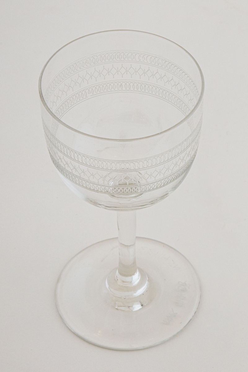 Rødvinsglass i klart glass, dekorert med etset og guillochert bord. Borden består av tre horisontale bånd med gjentagende geometriske figurer, i form av sirkel- og ruterformer. Klokkeformet kupa, hvor en ringformet vulst markerer overgangen til stetten. Sirkulær fotplate, noe hevet på midten.
