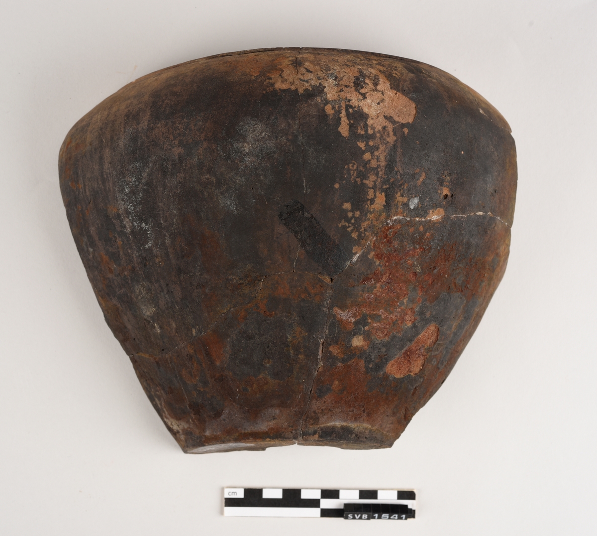 Krummet keramikkgjenstand bestående av fire fragment limt sammen. Innsiden er rødlig i farge med et sirkelformet mønstrer nederst på gjenstanden. Utsiden er svart i farge med rød/brune flekker.