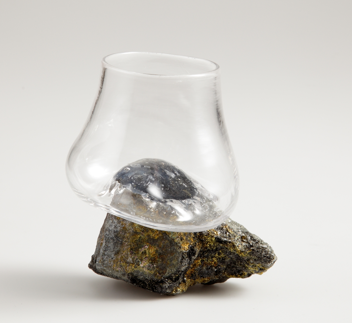 Dricksglas "On The Rocks" bestående av ett glas som är limmat på en sten. Glaset är friblåst och ser ut att ha sjunkit över den guld- och gråglimmande stenen.