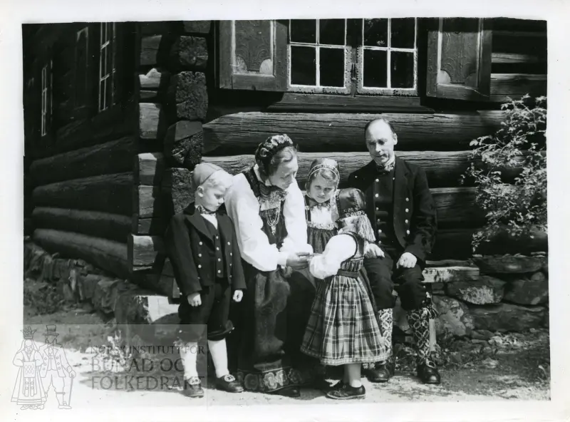 Kvinne, mann og barn i bunader foran tømmerhus.