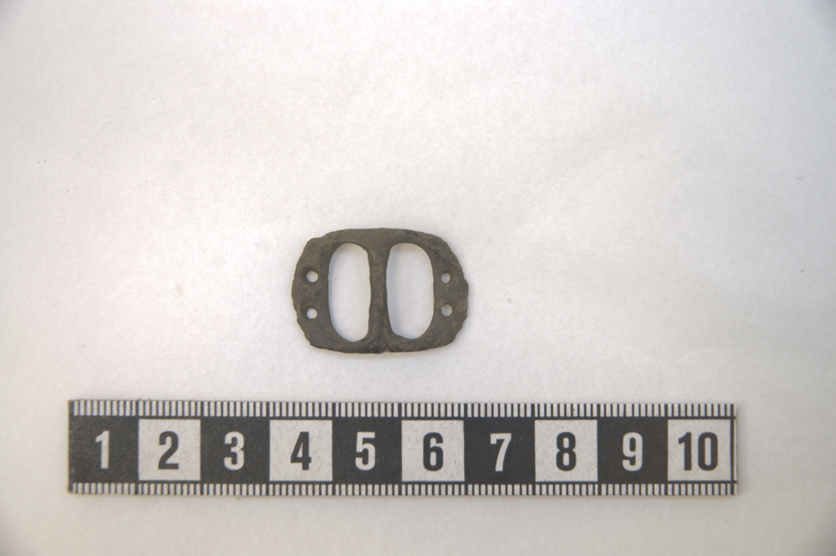 Remspenne av bronse, dobbeltspenne med oval spenneramme,  kanskje til sko. Spennen har to hull på de to ytre kantene. L: 27 mm, br.: 19 mm, t.: 4 mm. Vekt: 2,4 g.