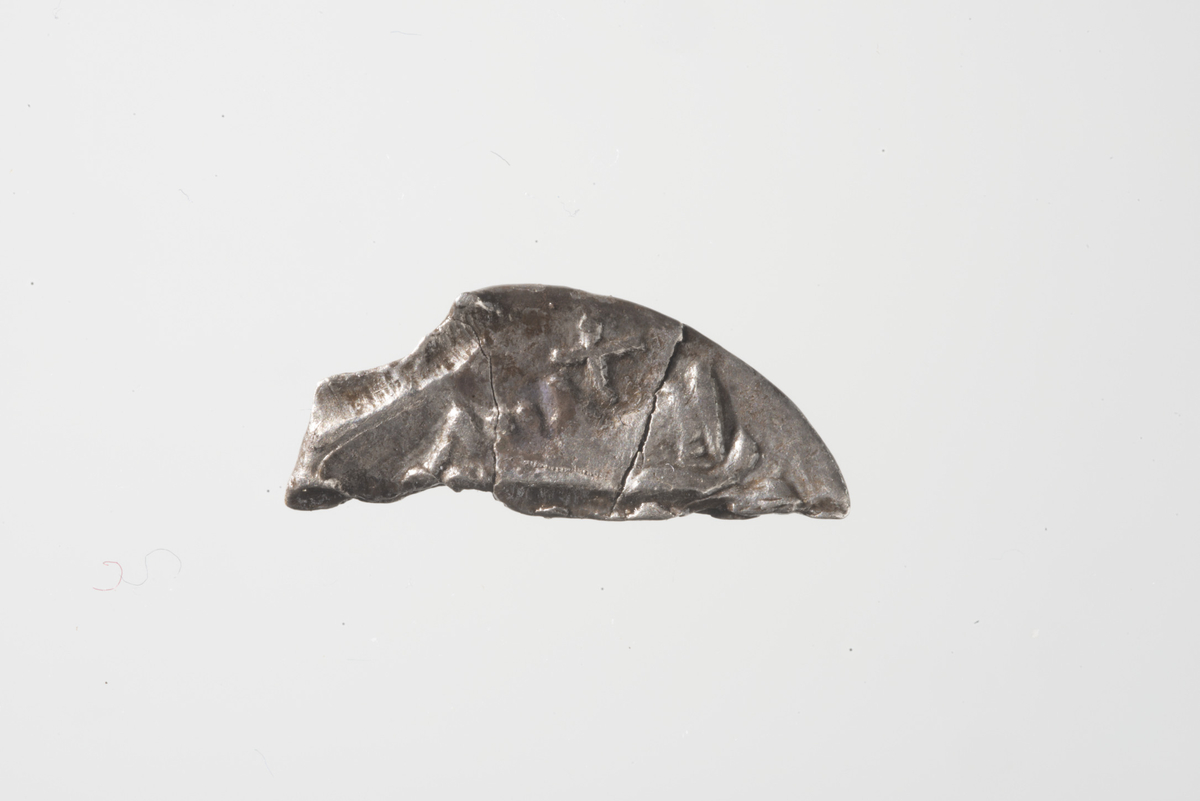 Mynt, trolig romersk sølvmynt, denar men vanskelig å identifisere. Mynten er knekt til 1/3 størrelse og er nedslitt. Advers: Nese, munn, hake og hals er synlig med et kors foran halsen. Ansikt vendt mot høyre. Revers: nedslitt overflate.