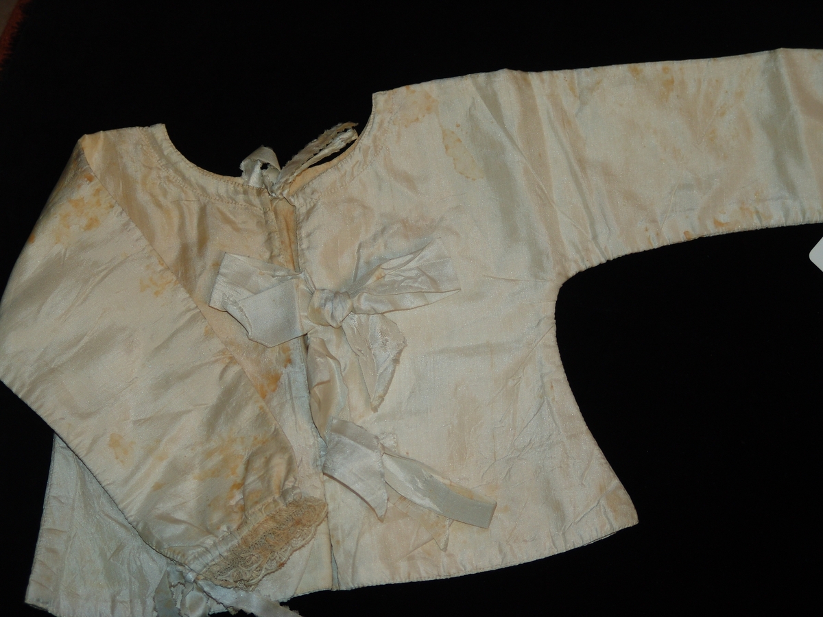 Liten barnskjorta tillverkad av vitt sidentyg. Snörning framtill i form av tre sidenband. Snörning i ärmarnas öppningar. Även spets i slutet på ärmarna.