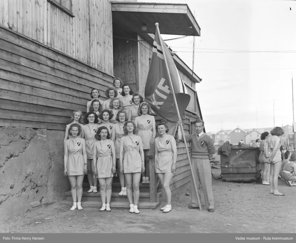 Turnstevne i Vadsø juni 1948. Kirkenes Idrettsforenings (KIF) dameturngruppe ved kinobrakka i Vadsø. 16 dameturnere og en mannlig fanebærer.. 