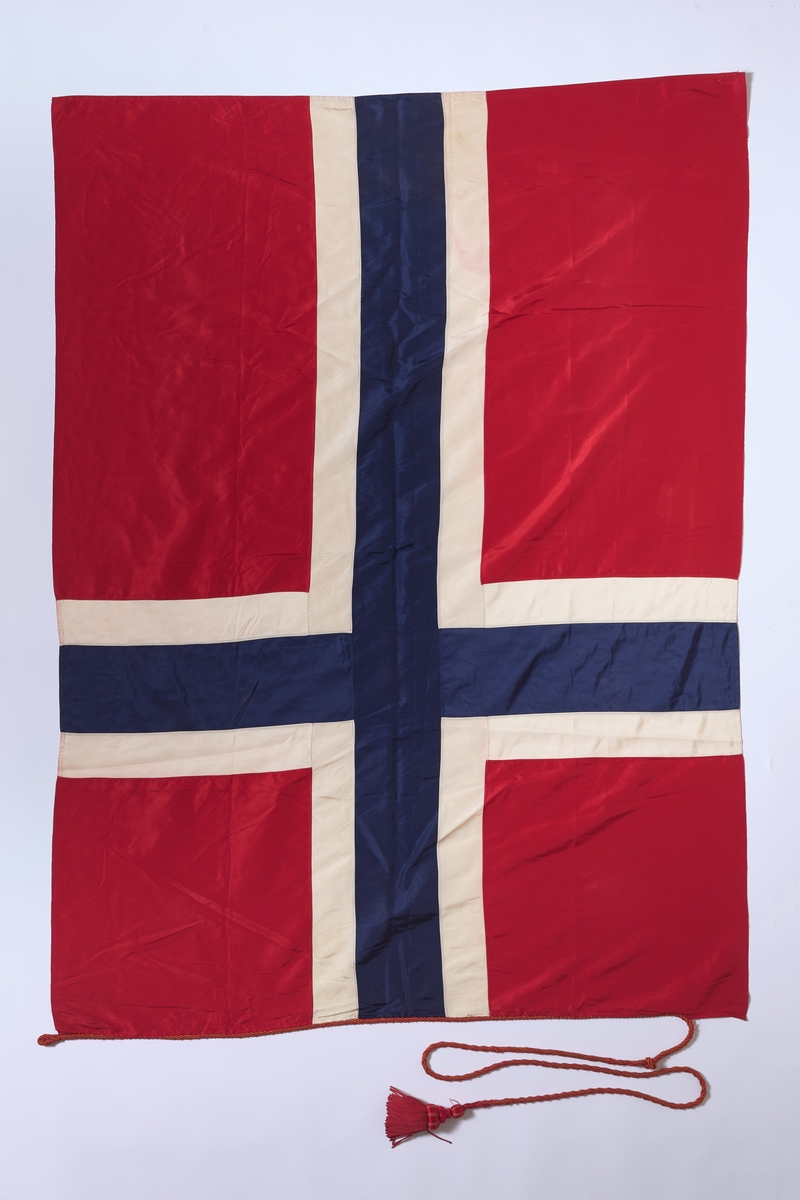 Norsk flagg sydd i kunstmateriale, med snor med dusk. 