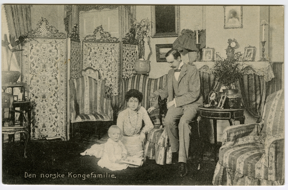 Postkort. Tekst på postkortet: "Den norske kongefamilie". Kong Haakon, dronning Maud og kronprins Olav sitter i en stue.