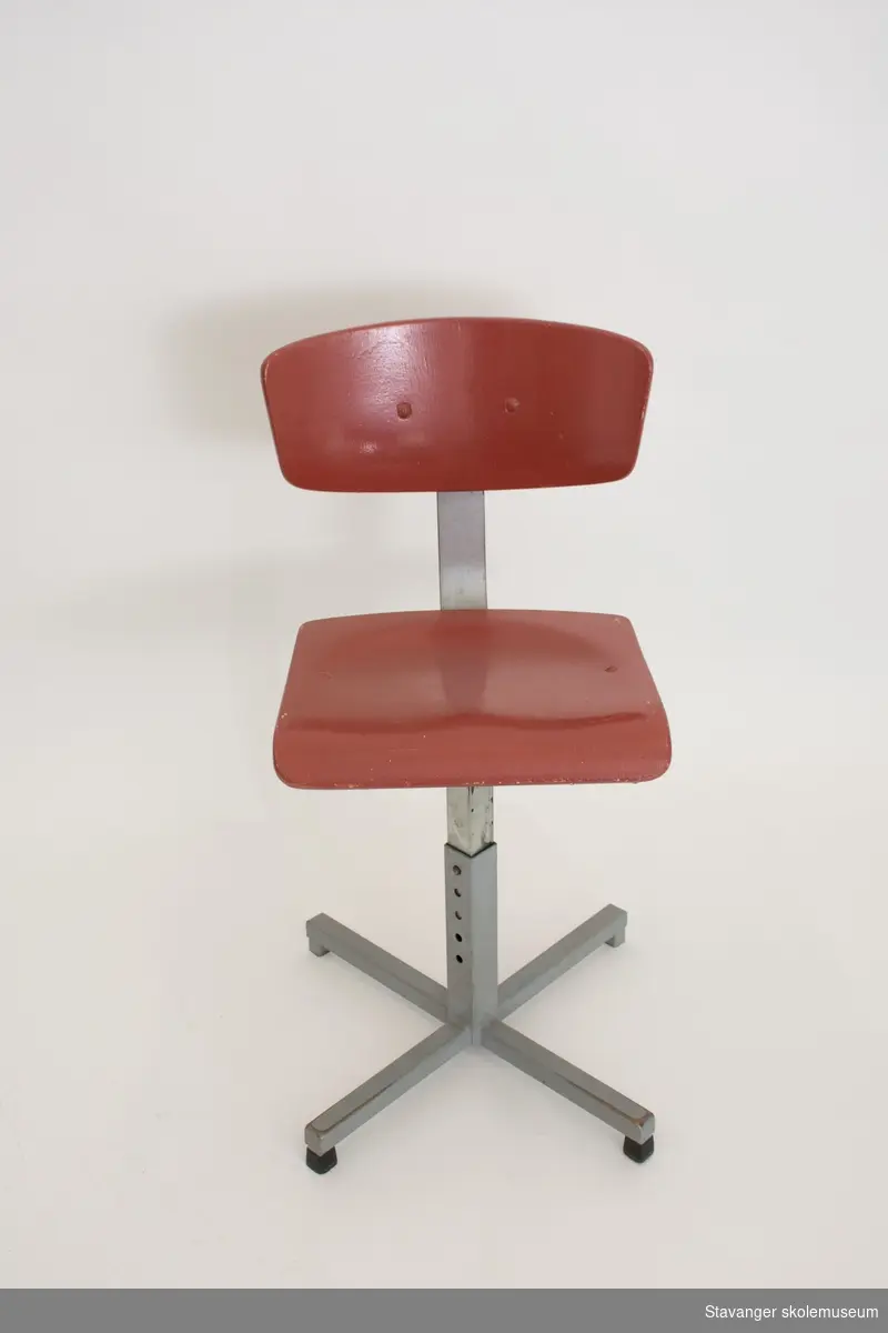 Elevstol av jern og bøyd kryssfiner. Gråmalt st]l og rødmalt kryssfiner i sete og rygg. Er justerbar i høyde.