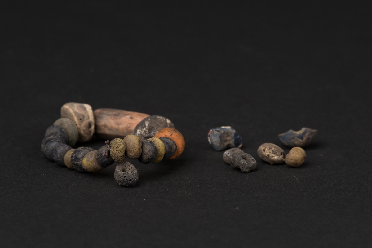 26 pärlor från ett gravfynd i olika material, storlek och form. 20 av pärlorna är trädda på en tråd.
De olika pärlorna är: en lång, åttkantig karneolpärla, en orangegul tunnformig glasflusspärla, två hela och två halva blå glaspärlor med insatta, olikfärgade trådar eller ögon (de hela är eldskadade) samt 20 små, ringformade glaspärlor av mörkblå eller citrongul färg, några av dessa är starkt eldskadade.