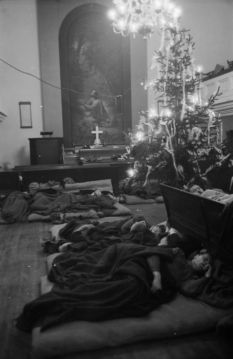 Personer sover i Metodistkirkenen i Trondheim i jula 1944. De ligger på madrasser på gulvet og på kirkebenker. De har enkle pledd å dekke seg til med. I bakgrunnen ser man prekestolen, alteret med altertavle, juletreet og galleriet.