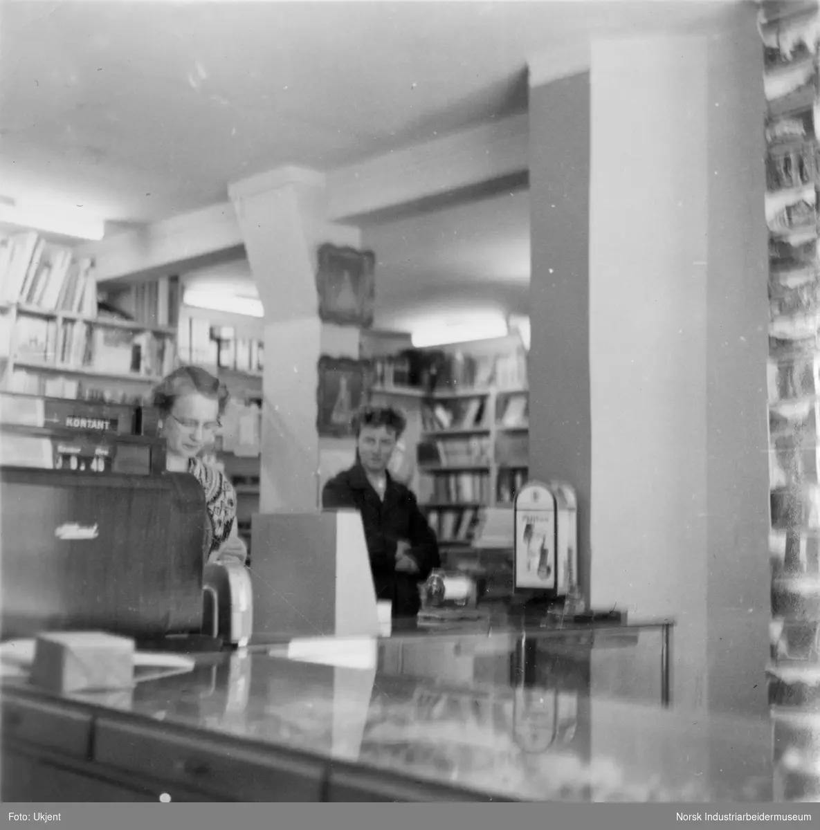 Avdeling hos Køhns bokhandel og trykkeri. To damer står bak disken med kassaapparat. Bøker i hyllene på baksiden.