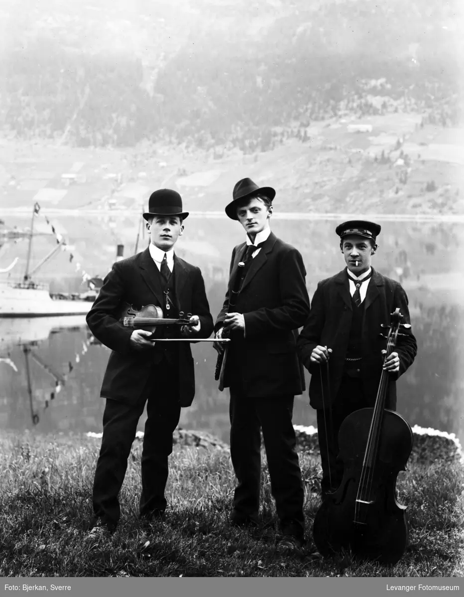 Gruppebilde av tre menn med cello og fioliner, tatt ue ved vann/fjord, båt i bakgrunnen