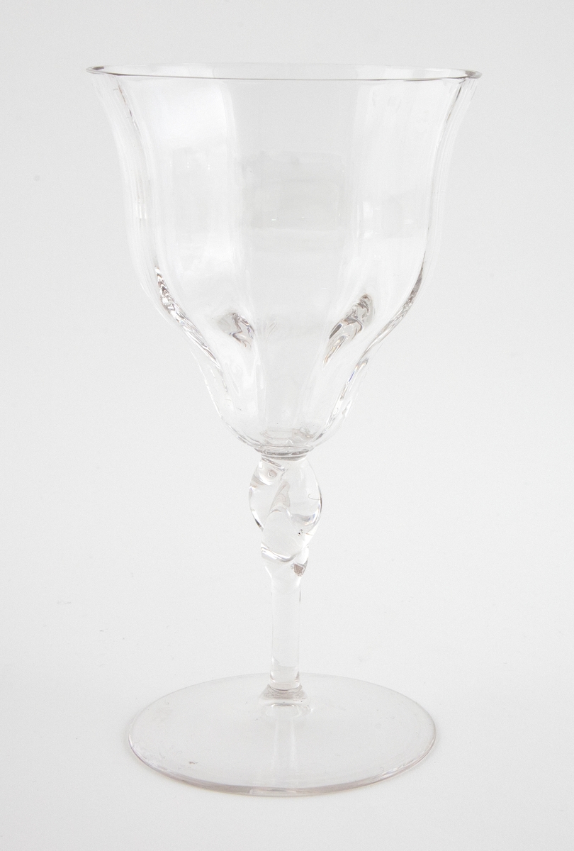 Vannglass i klart glass med vertikale riller. Konisk kupa med bølgende utforming, som hviler på en høy og slank stett. Overgangen mellom kupa og stett er markert med en knuteformet vulst.