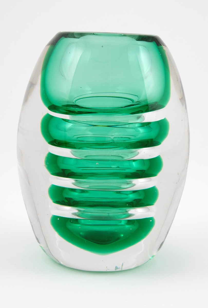Ovalformet vase i underfangsglass. Et indre sjikt i grønt gjennomskinnelig glass, som er utformet som ringformede vulster, omgitt av et tykt lag av klart glass. Sirkulær åpning og planslipt bunn.