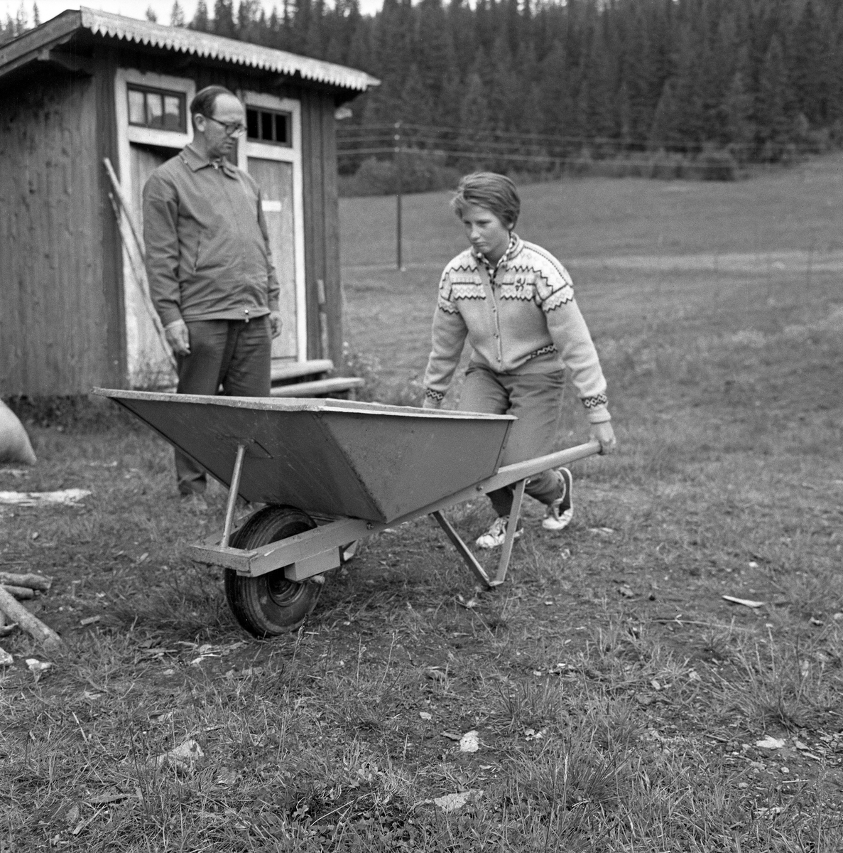 Jente med trillebår, fotografert under en 4H-leir i Søre Osen i Trysil i Hedmark i 1962. Da dette bildet ble tatt sto jenta med bøyde knær, enten for å løfte eller sette fra seg den tunge båra. En voksen mann sto og betraktet henne, antakelig for å bedømme arbeidet. Bak han ser vi en utedo med to rom.