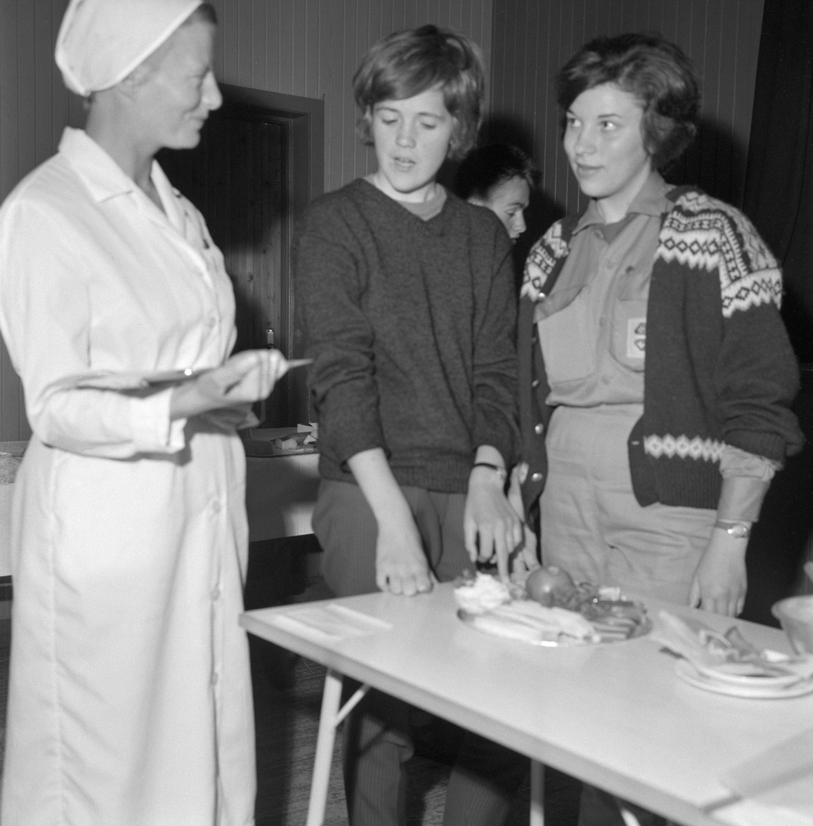 Presentasjon av 4H-prosjekt undr 4H-leir i Søre Osen i Trysil i Hedmark i 1962. Vi ser tre kvinner framfor et respetexbord, der det sto et ostefat. Sannsynligvis var det en den unge kvinna i midten som hadde anrettet osten. Den kvitkledde kvinna til venstre var sannsynligvis en fagperson, muligens en husstellærer, som skulle vurdere arbeidet. Kvinna til høyre hadde et 4H-emblem på blusen, delvis dekt av strikkejakka. Det er nærliggende å anta at hun representerte organisasjonen.