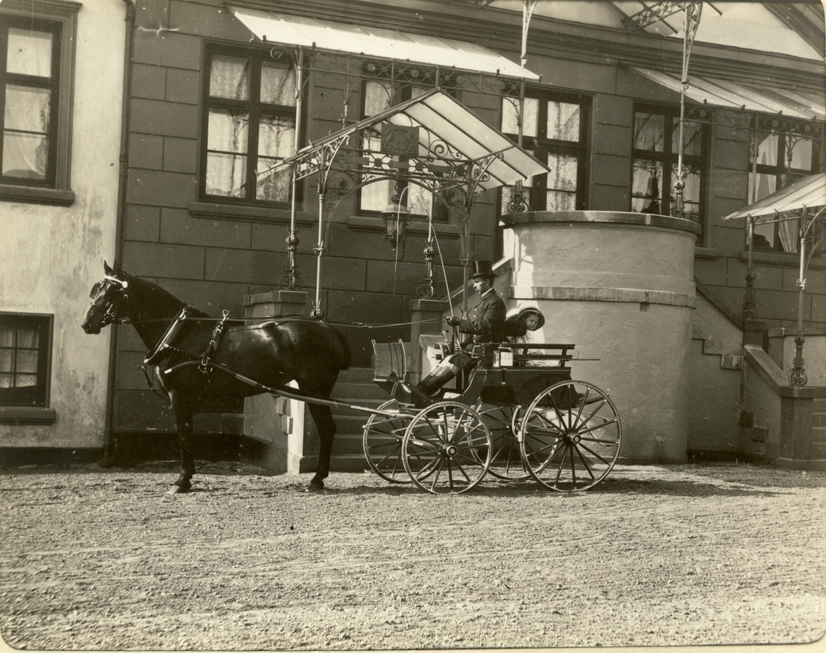 Hesten «Coquette» er spent for vognen. Kusk og passasjer, én av søstrene Egeberg, er parat til avgang på gårdsplassen til Bogstad gård. Fotografert 1911.