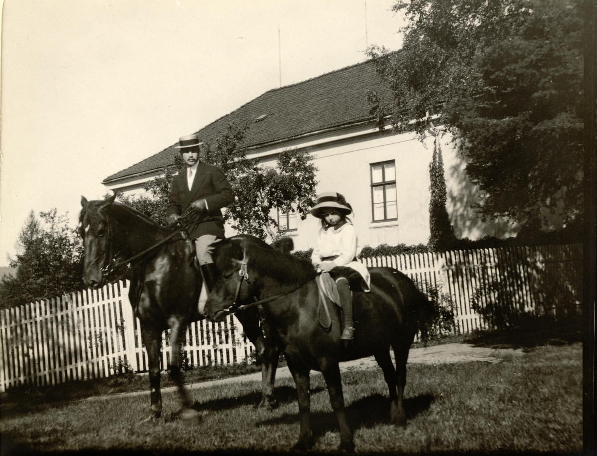 Westye Egeberg på hesten "Shorthand" og datteren Mimi på hesten "Dokka" foran en hovedbygning, antagelig Skinnatbøl i Kongsvinger. Fotografert 1911.