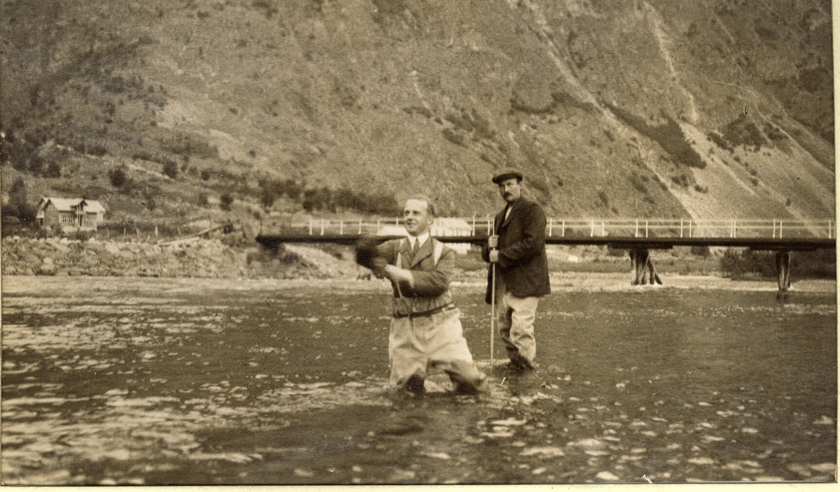 Thorleif Tjersland fisker i Lærdalselvi, Sogn og Fjordane. Bak står en medhjelper (Lasse?) med klepp. I bakgrunnen en bro over elven og til venstre et våningshus. Fotografert sommeren 1916.