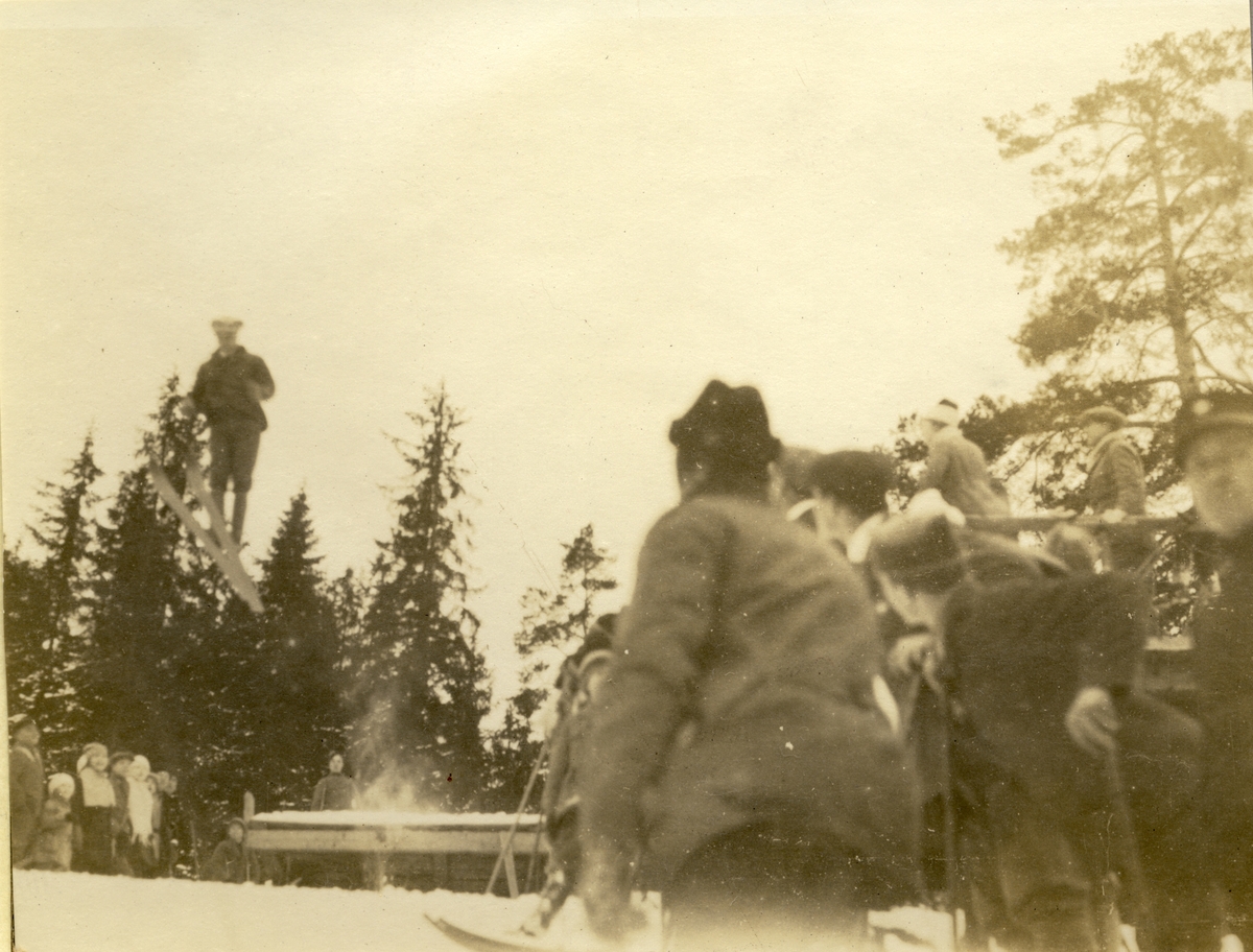 Einar, antagelig Einar Westye Egeberg, hopper på ski fra Tangenbakken i Sørkedalen. Tilskuere og dommere, øverst til høyre, følger med. Fotografert 1918.