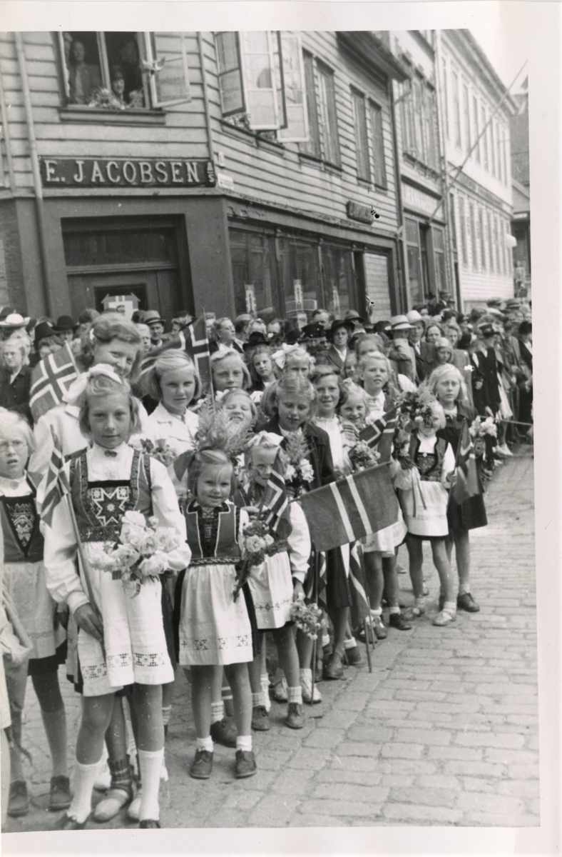 Blomsterpiker i finstasen i bakken ved Tekstilhandel E. Jacobsen i Strandgt, som venter på kronprins Olav som besøkte Haugesund Olsokdagen 29. juli 1945.