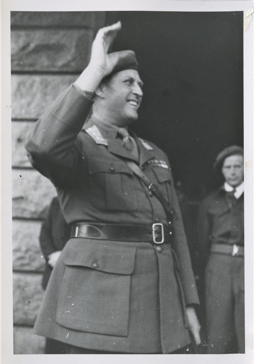 Norges Forsvarssjef kronprins Olav ankommer Haugesund Olsokdagen 29. juli 1945. Her ser vi han stå på trappen til Rådhuset vinkende og smilende til de fremmøtte på Rådhusplassen. Bak til høyre ser vi Hjemmestyrkenes distriksjef Alf Skare